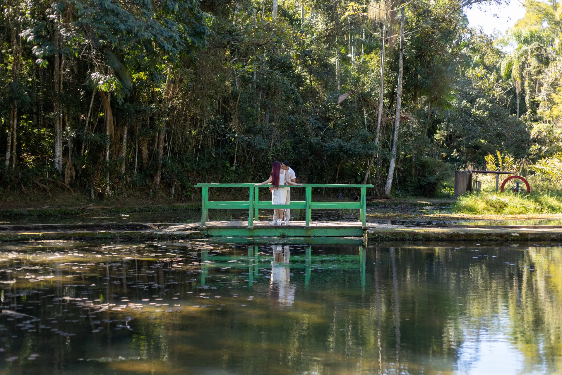 Ketelyn e Carlos se beijando em uma ponte verde à beira do lago cercado pela mata